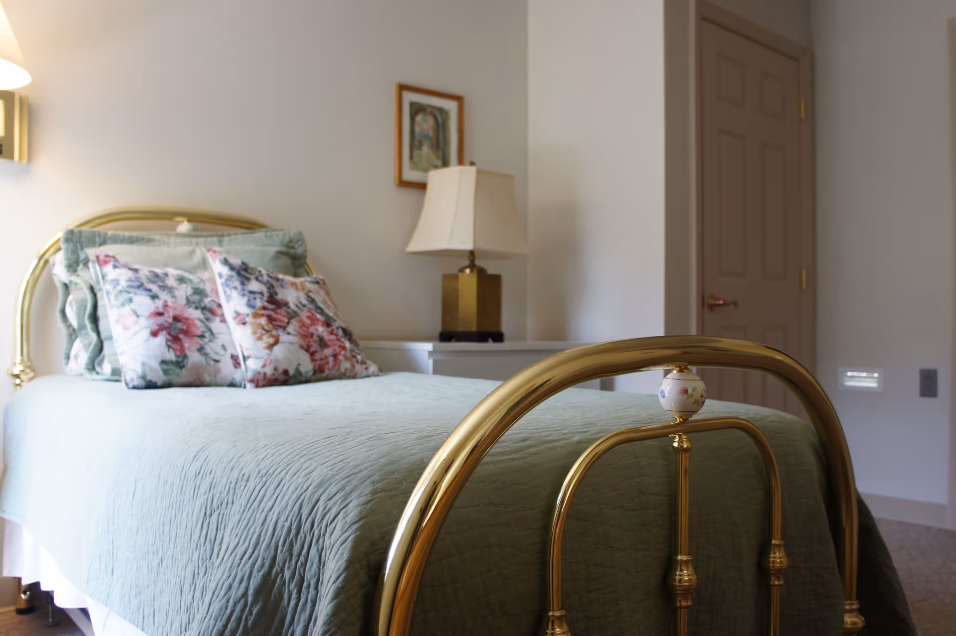 A bedroom with a brass bed frame, a light green quilted bedspread, and floral pillows. There is a white nightstand with a brass and white lampshade lamp, a framed picture on the wall, and a closed beige door in the background.