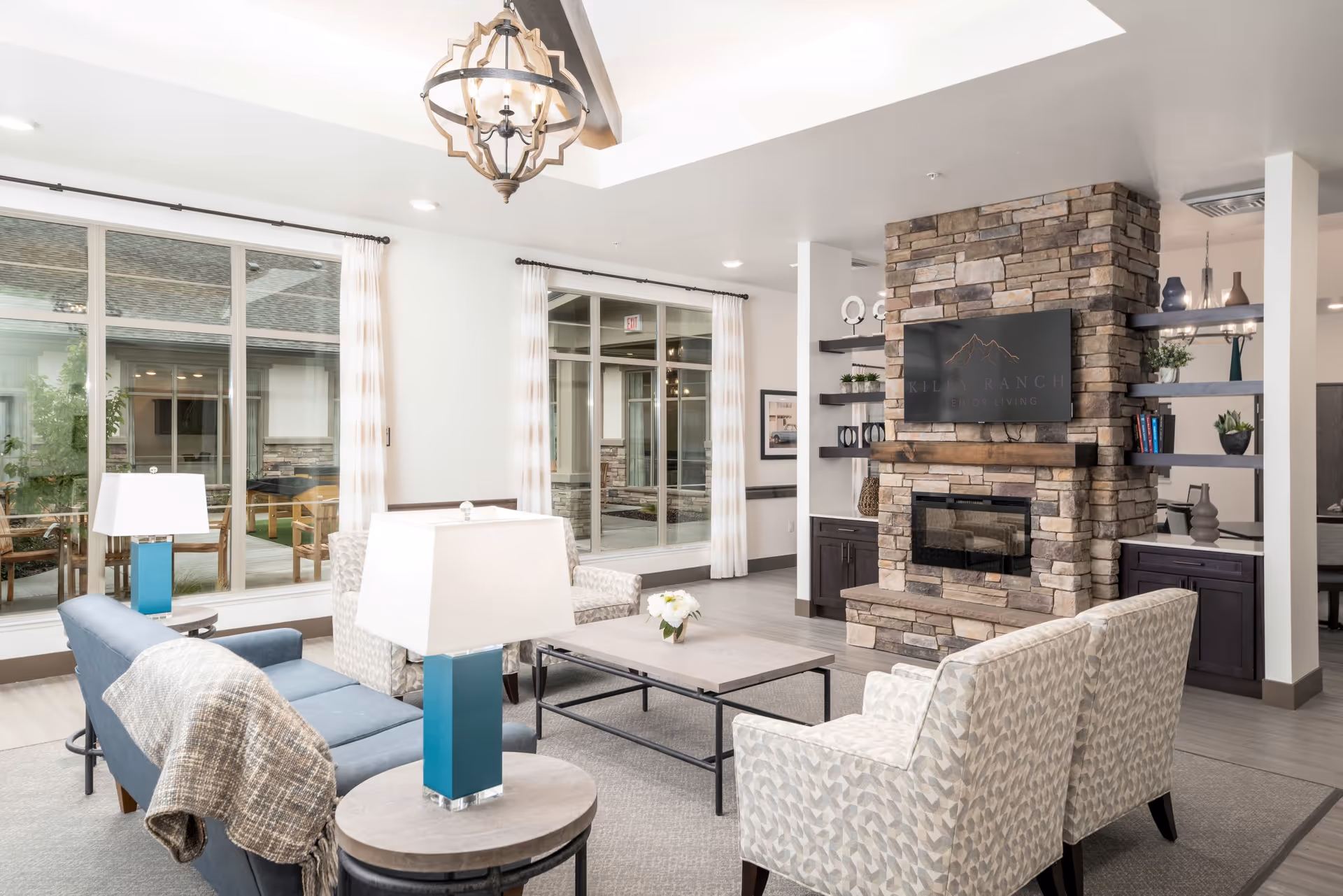 A bright and spacious senior living facility common area with large windows, light curtains, a stone fireplace with a mounted TV displaying the Kiley Ranch Senior Living logo, comfortable seating including a blue sofa and patterned armchairs, a wooden coffee table with a small flower arrangement, and modern lamps on side tables.