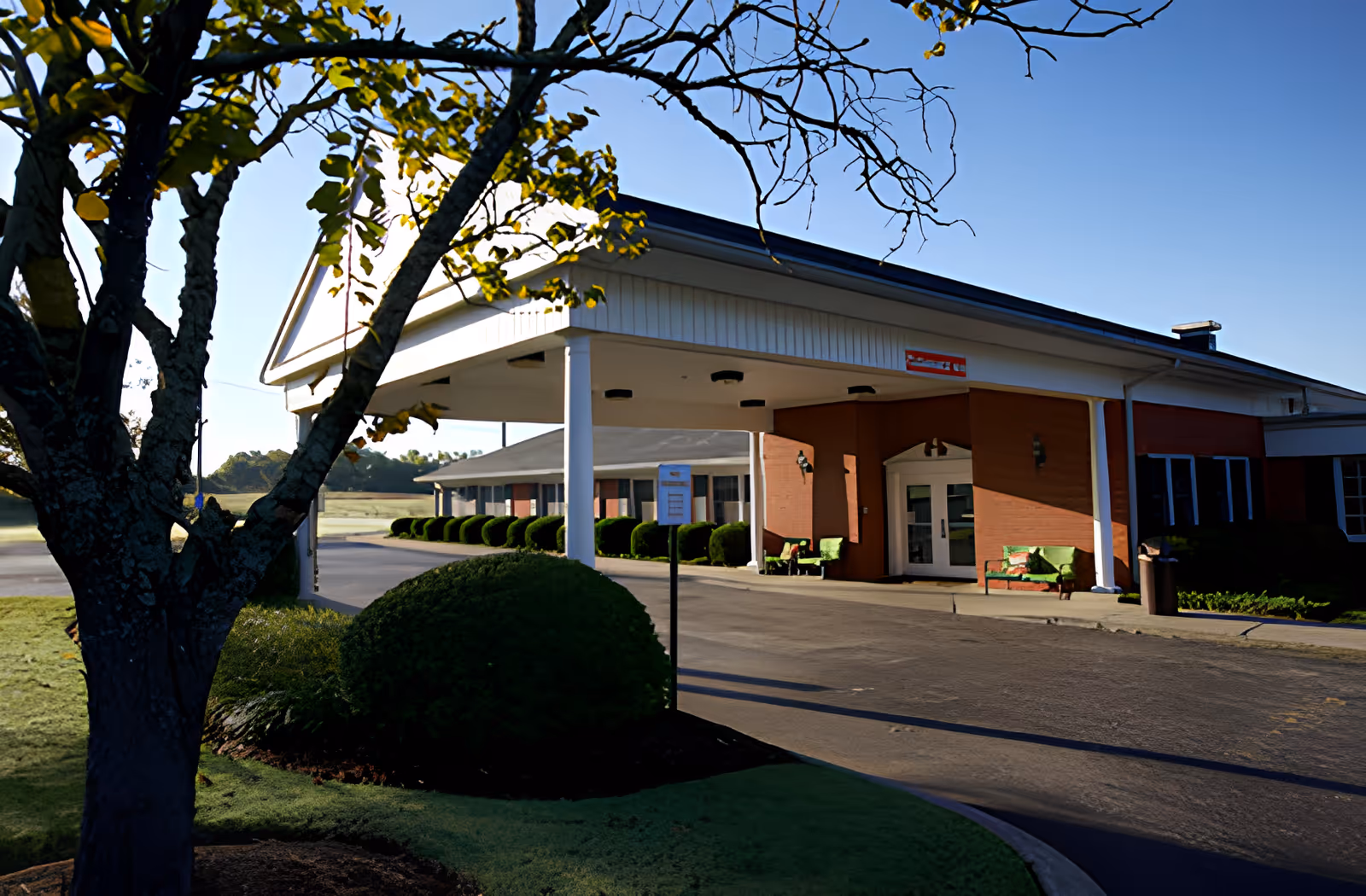 Exterior view of a single-story building with a covered entrance supported by white columns. There are green bushes and a tree in the foreground, and two green benches near the entrance. The sky is clear and blue.