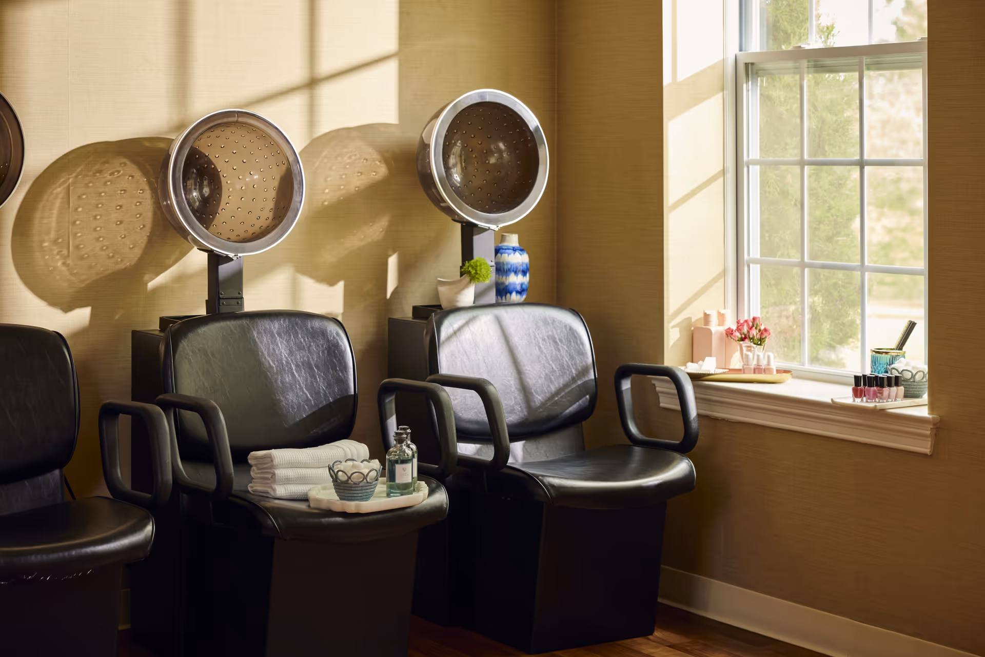 Interior of a salon area with two black leather salon chairs under hair dryers, a window with sunlight streaming in, and a windowsill holding nail polish bottles and other manicure supplies.