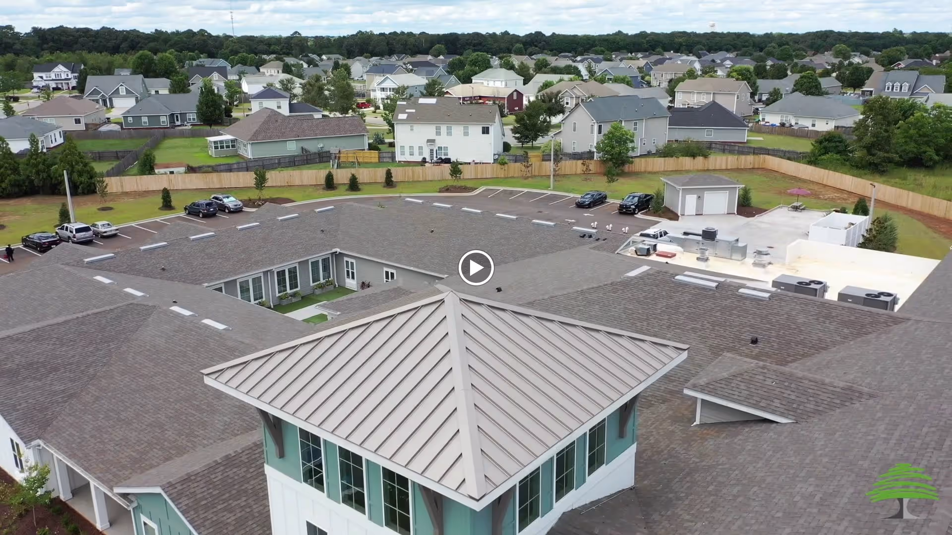 Aerial view of The Canopy at Warner Robins senior living facility showing the building's roof, surrounding parking lot with several cars, and nearby residential neighborhood with houses and trees under a cloudy sky.