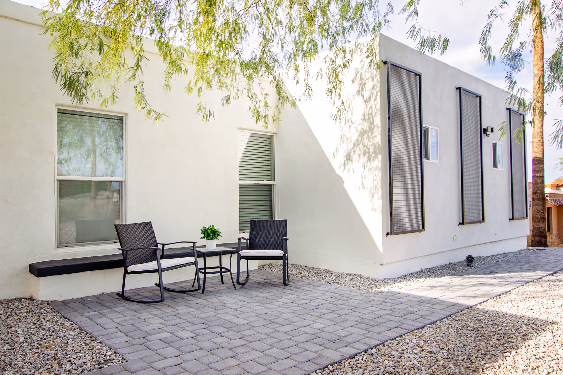Outdoor patio area with two black chairs and a small table with a potted plant between them, set on a paved stone surface next to a white building with windows and decorative panels. Trees cast shadows on the building and ground.