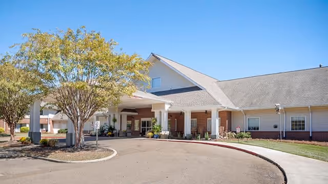 Exterior view of a single-story senior living facility building with a covered entrance, a circular driveway, and a tree with green leaves in front under a clear blue sky.