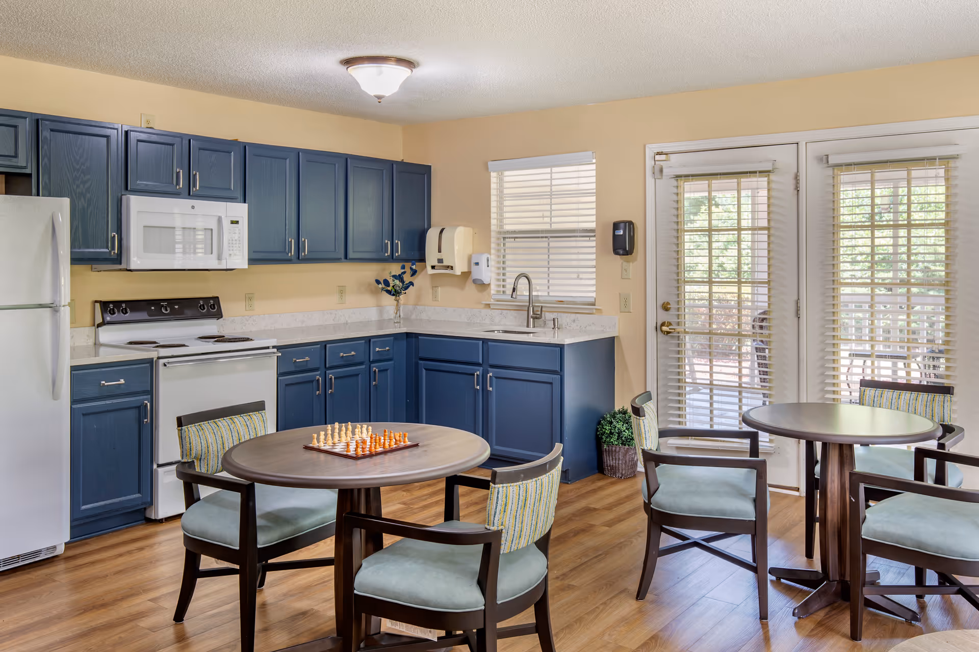 A communal kitchen and dining area with blue cabinets, white appliances, round tables and chairs, and French doors to the outside.