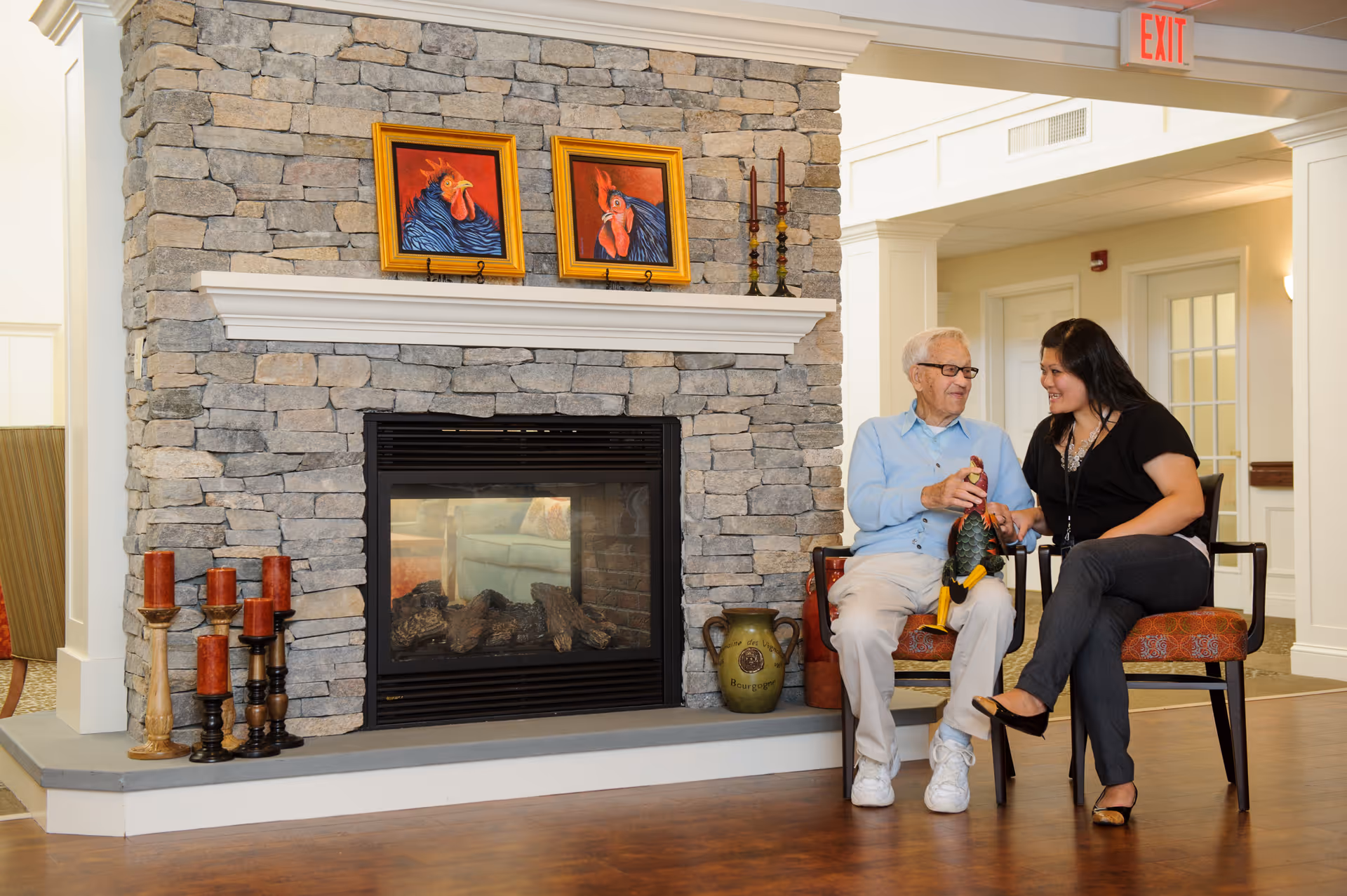 An elderly man and a woman sitting on chairs in a cozy room with a stone fireplace. The man is holding a decorative rooster figurine while the woman smiles and interacts with him. The fireplace mantel has two framed rooster paintings and several candle holders. The room has warm lighting and a wooden floor.