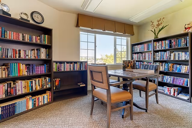 A bright room with large windows showing an outdoor view, featuring multiple bookshelves filled with books along the walls. In the center, there is a table with four chairs around it, creating a cozy reading or gathering space.