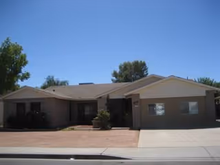 Single-story beige-brick assisted living building with a driveway and front yard under blue sky.