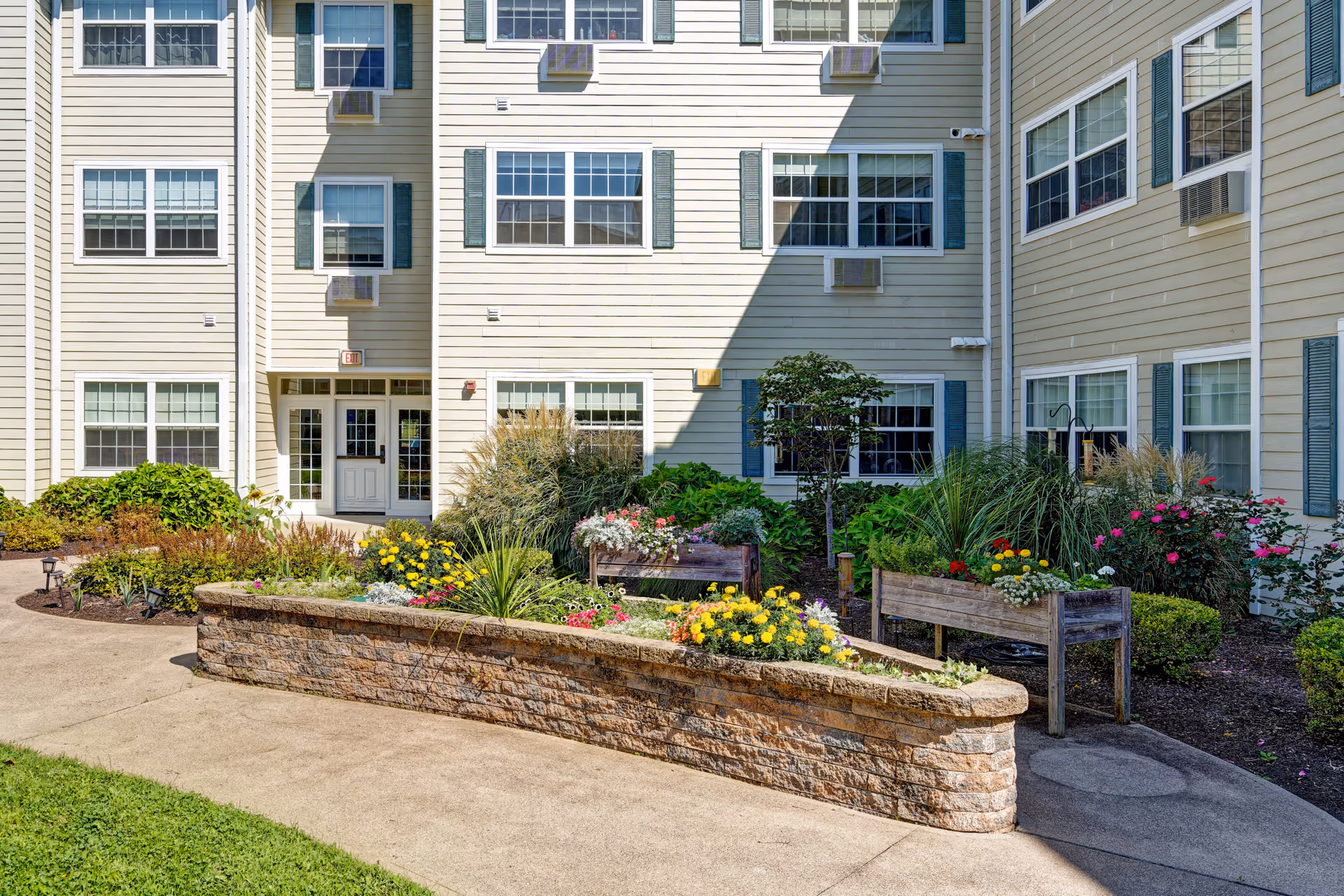 Sunlit landscaped courtyard with raised flowerbeds and benches in front of a multi-story light-colored senior living building with many windows and a central entrance.