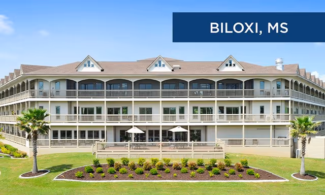 Exterior view of a large, three-story building with multiple balconies and a landscaped garden in front, located in Biloxi, Mississippi. The building has a light-colored facade and a sloped roof with dormer windows.