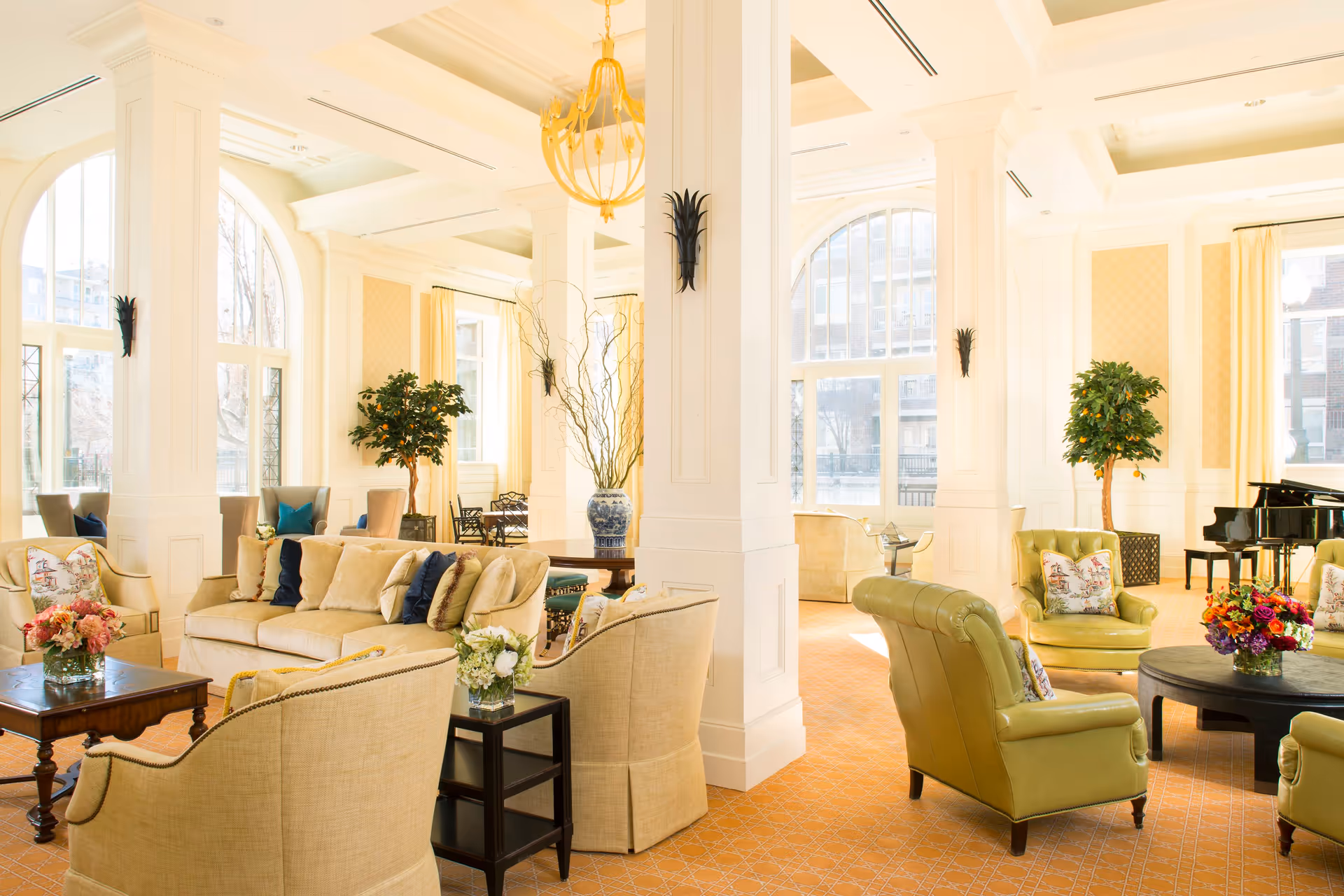 Bright and spacious senior living facility lounge area with large arched windows letting in natural light. The room features comfortable beige and green upholstered chairs and sofas arranged around wooden coffee tables with floral centerpieces. Tall white columns, potted plants, and a grand piano add elegance to the space.