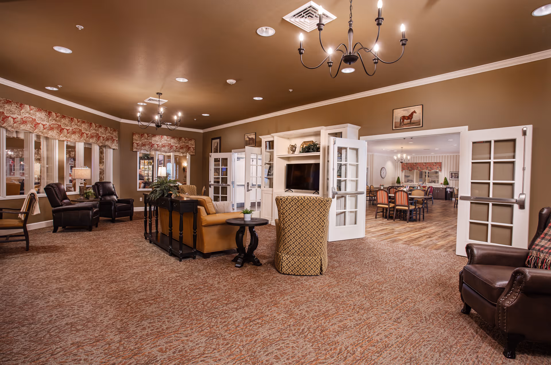 A spacious senior living facility common area with comfortable seating including leather and upholstered chairs, a tan sofa, and a small round table. The room has brown walls, carpeted flooring, and two chandeliers hanging from the ceiling. French doors lead to an adjacent dining area with wooden flooring and multiple dining tables and chairs. The walls are decorated with framed pictures, including one of a horse above the French doors.