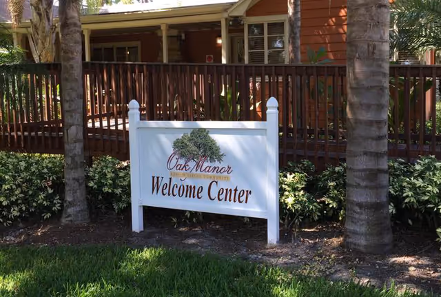 White sign reading 'Oak Manor Welcome Center' in a landscaped yard with palm trees and a wooden building behind it.