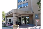 Entrance of Elm York Assisted Living facility showing a covered drop-off area with columns, glass doors, and a brick exterior. There are flower pots and a person sitting on a bench near the entrance.