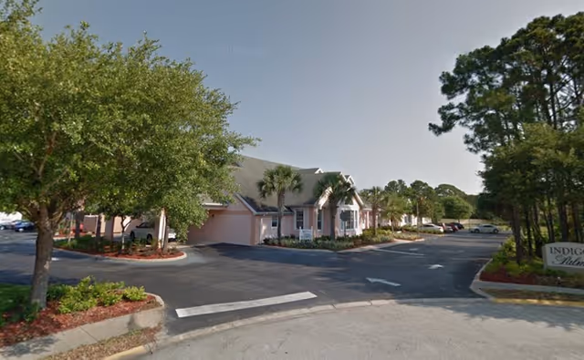 Exterior view of Indigo Palms facility showing a single-story building with a light pink exterior, surrounded by palm trees and other greenery. There is a paved parking lot with several parked cars and a sign that reads 'Indigo Palms' near the entrance.