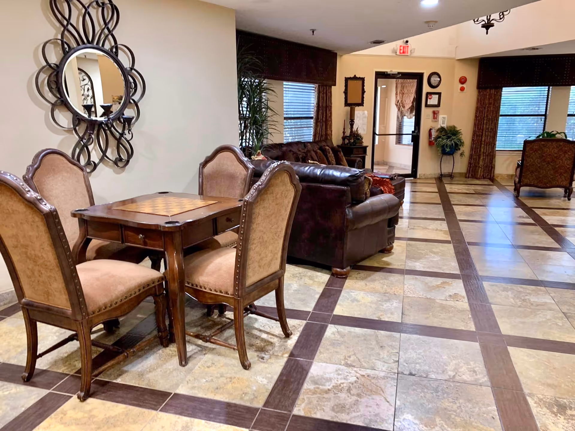 A cozy interior common area with a small wooden table and four upholstered chairs in the foreground. Behind the table is a dark brown leather sofa and additional seating near windows with curtains. The floor is tiled with a pattern of large square tiles and dark brown borders. A decorative round mirror with a black ornate frame hangs on the wall to the left. There is a door with a glass panel and an exit sign above it in the background.