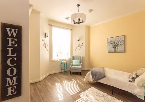 Sunny, softly decorated single-occupancy bedroom with a bed, armchair by a window, small side table, and a vertical "WELCOME" sign on the wall.