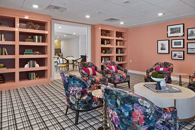 Bright communal lounge with floral-upholstered armchairs around a round coffee table, pink built-in bookshelves, framed photos on coral walls and a view into an adjoining dining area.