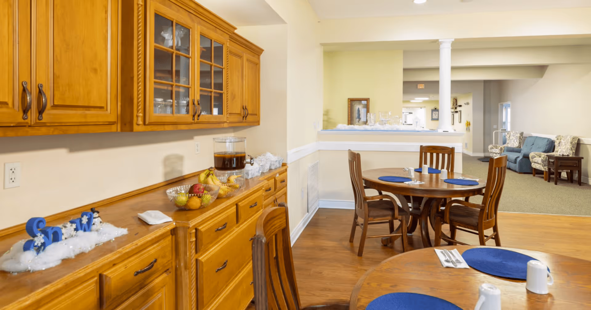 Dining area in an assisted living facility with wooden tables and chairs set with blue placemats, white mugs, and silverware. A wooden cabinet with glass doors holds dishes and glassware, and a countertop displays a beverage dispenser and bowls of fruit. In the background, there is a seating area with upholstered chairs and a small table.
