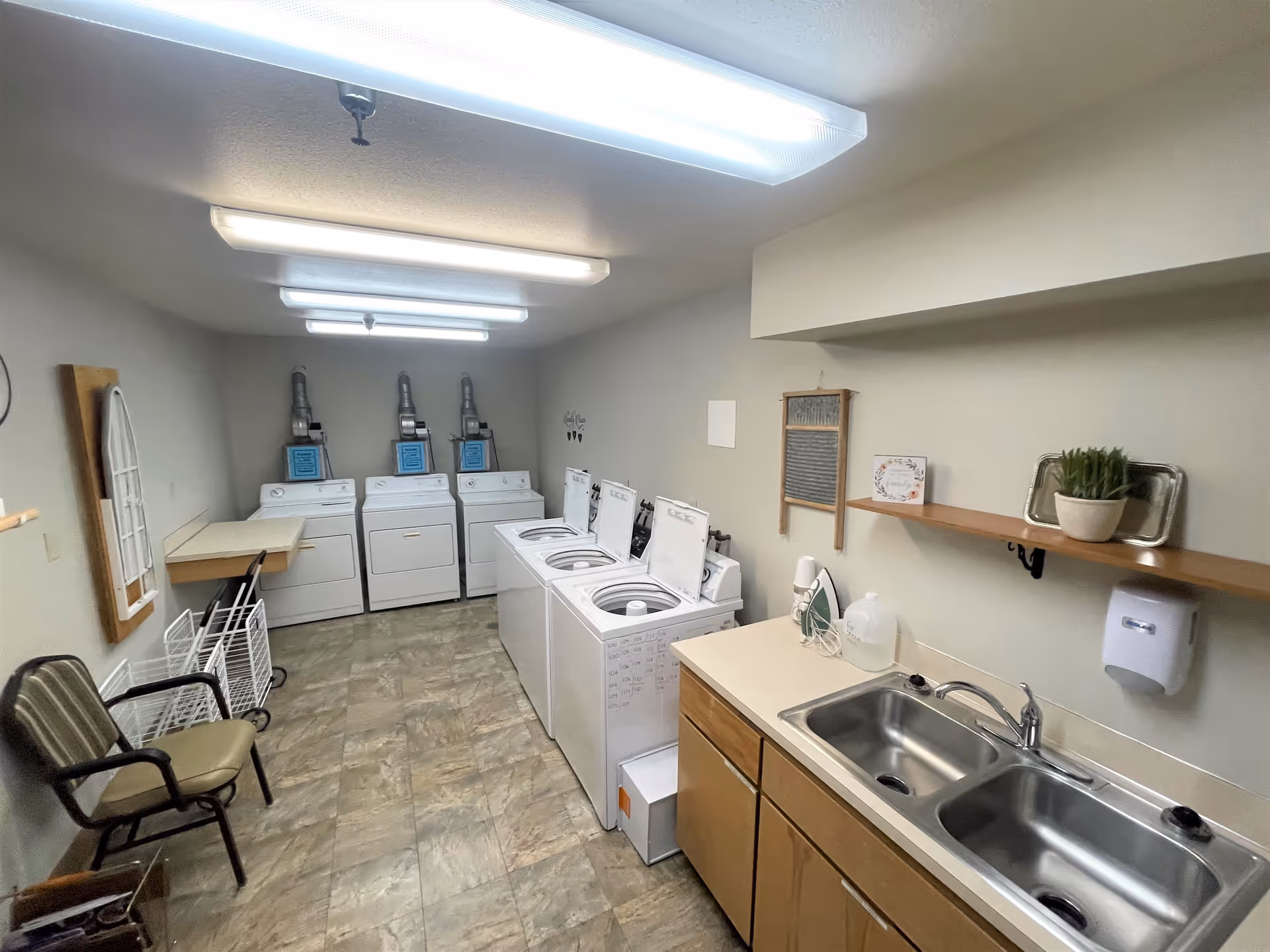 Laundry room with three top-loading washing machines and three front-loading dryers along the walls. There is a double sink with a countertop and cabinets underneath on the right side. A chair, folding table, and laundry carts are on the left side. The room is well-lit with fluorescent ceiling lights and has a tiled floor.