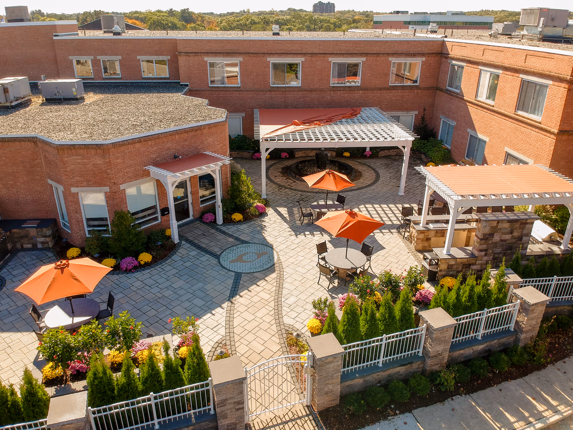 Aerial view of an outdoor courtyard at a senior living facility with brick buildings surrounding it. The courtyard features paved walkways, several round tables with orange umbrellas, a white pergola, a stone outdoor kitchen or barbecue area with a pergola, and landscaped greenery and flowers along the edges. There is a gated entrance with a white fence and stone pillars.