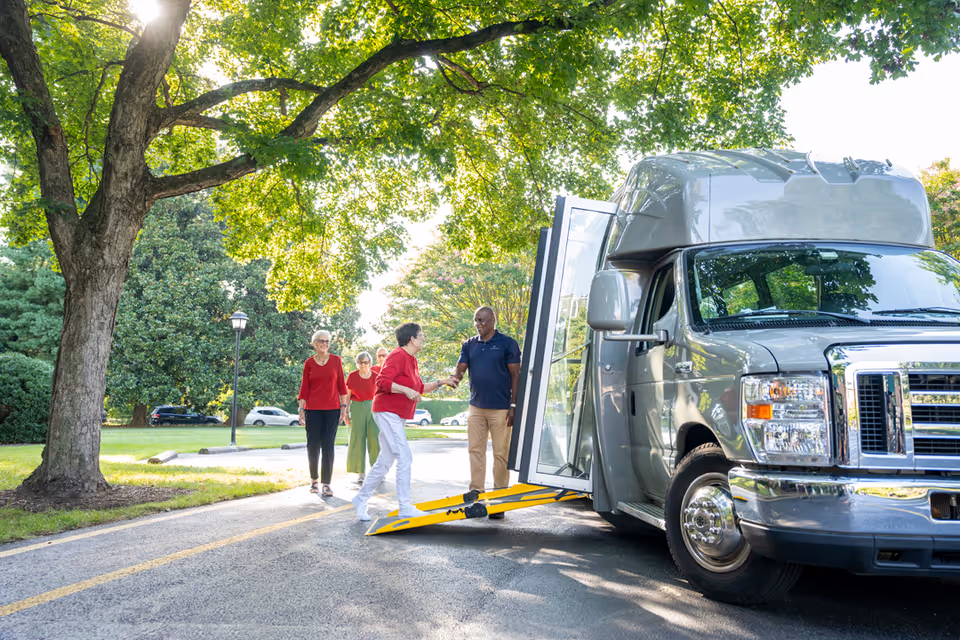 A group of elderly people outdoors near a large vehicle with an extended ramp. One elderly woman is shaking hands with a man standing by the vehicle door, while two other elderly women walk towards them on a paved path under a large tree with green leaves.