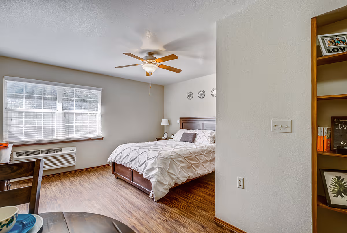 A bright bedroom with a large window covered by white blinds, a ceiling fan with wooden blades, a wooden bed with white bedding and a gray pillow, a nightstand with a lamp and clock, and a wooden bookshelf partially visible on the right side.