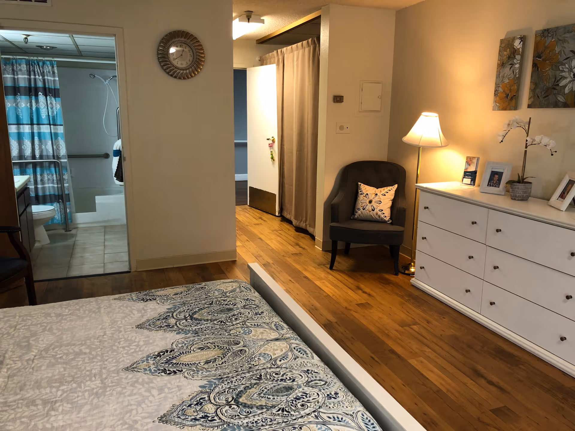 Bedroom with a patterned bedspread in the foreground, a white dresser and armchair with a lamp, and an open doorway showing a bathroom.