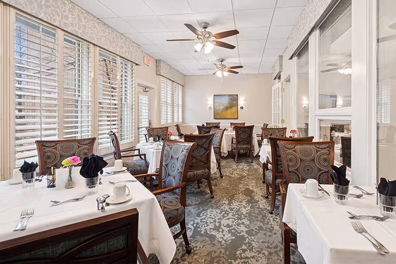 A bright dining room with several tables covered in white tablecloths, each set with cups, utensils, and napkins. The chairs have patterned upholstery, and there are large windows with white blinds letting in natural light. Ceiling fans with lights hang from the ceiling, and a painting is visible on the far wall.