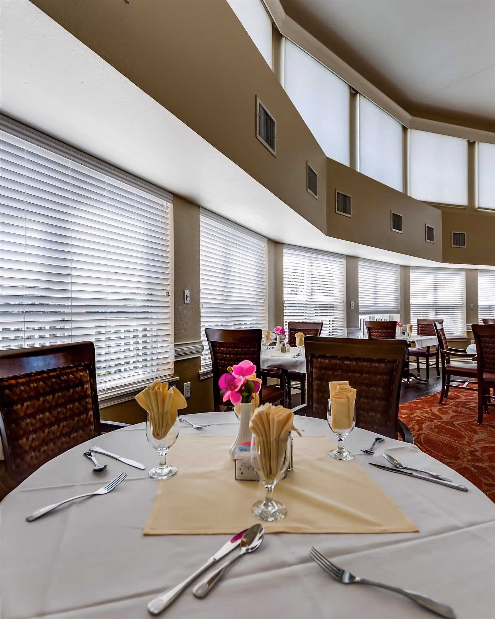 Sunlit dining room with round tables set with napkins, silverware, and small floral centerpieces beside large windows with blinds.