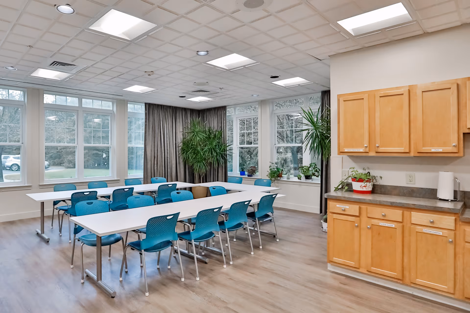 A bright, spacious room with large windows letting in natural light. The room features two long white tables arranged in a U-shape with blue chairs around them. There are several green plants near the windows and a set of wooden cabinets with a countertop on the right side. The floor is light wood, and the ceiling has recessed lighting and a grid pattern.