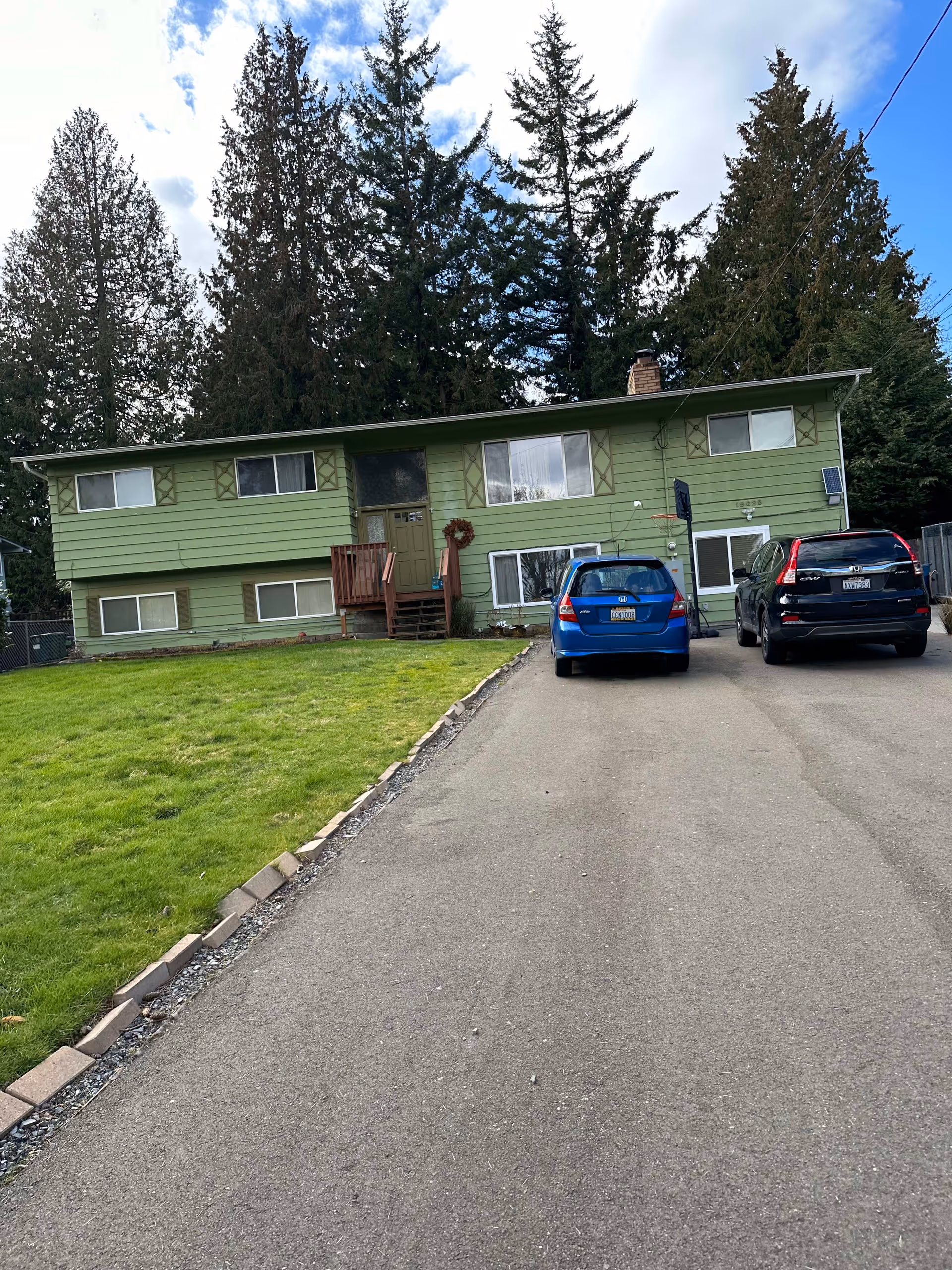Front exterior view of a green two-story house with a driveway and two parked cars, surrounded by tall evergreen trees under a partly cloudy sky.