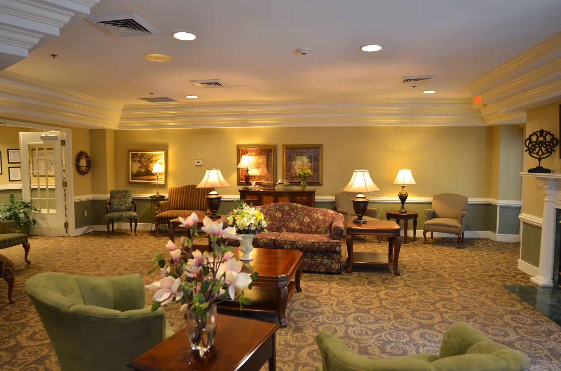 Well-lit furnished lounge with sofas, armchairs, coffee tables, lamps, and floral arrangements.