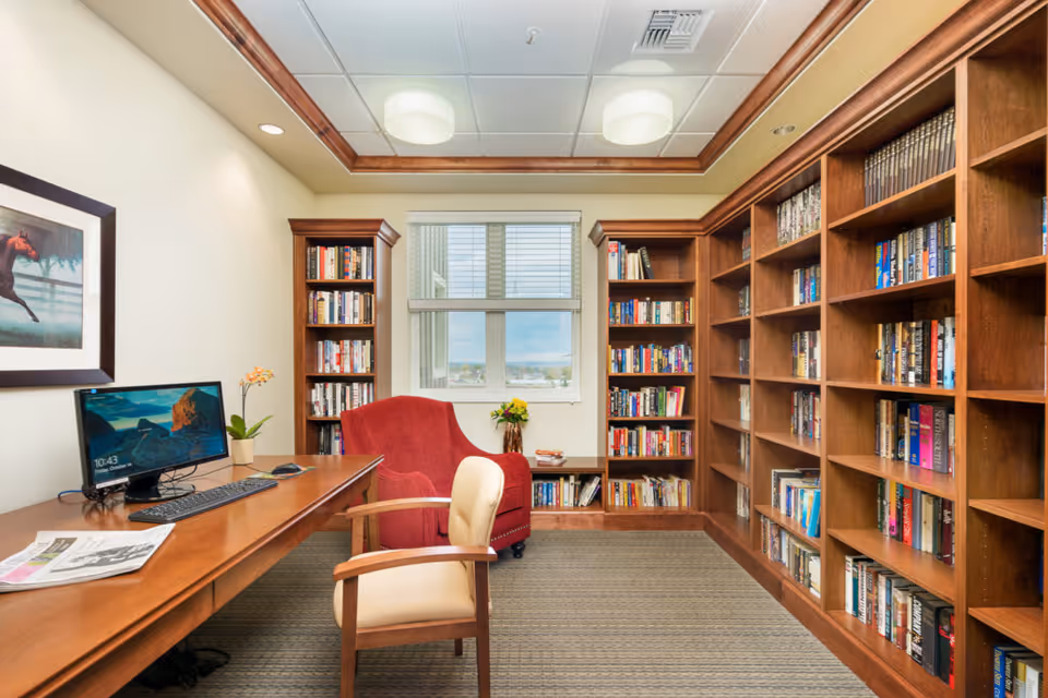 A cozy library room with wooden bookshelves filled with books along the right wall and back wall. There is a large wooden desk with a computer, keyboard, mouse, and a newspaper on it. Two chairs are present: a red upholstered armchair near the window and a beige cushioned chair in front of the desk. A framed picture of a horse hangs on the left wall. The ceiling has recessed lighting and a wooden trim. A window in the center back wall lets in natural light.