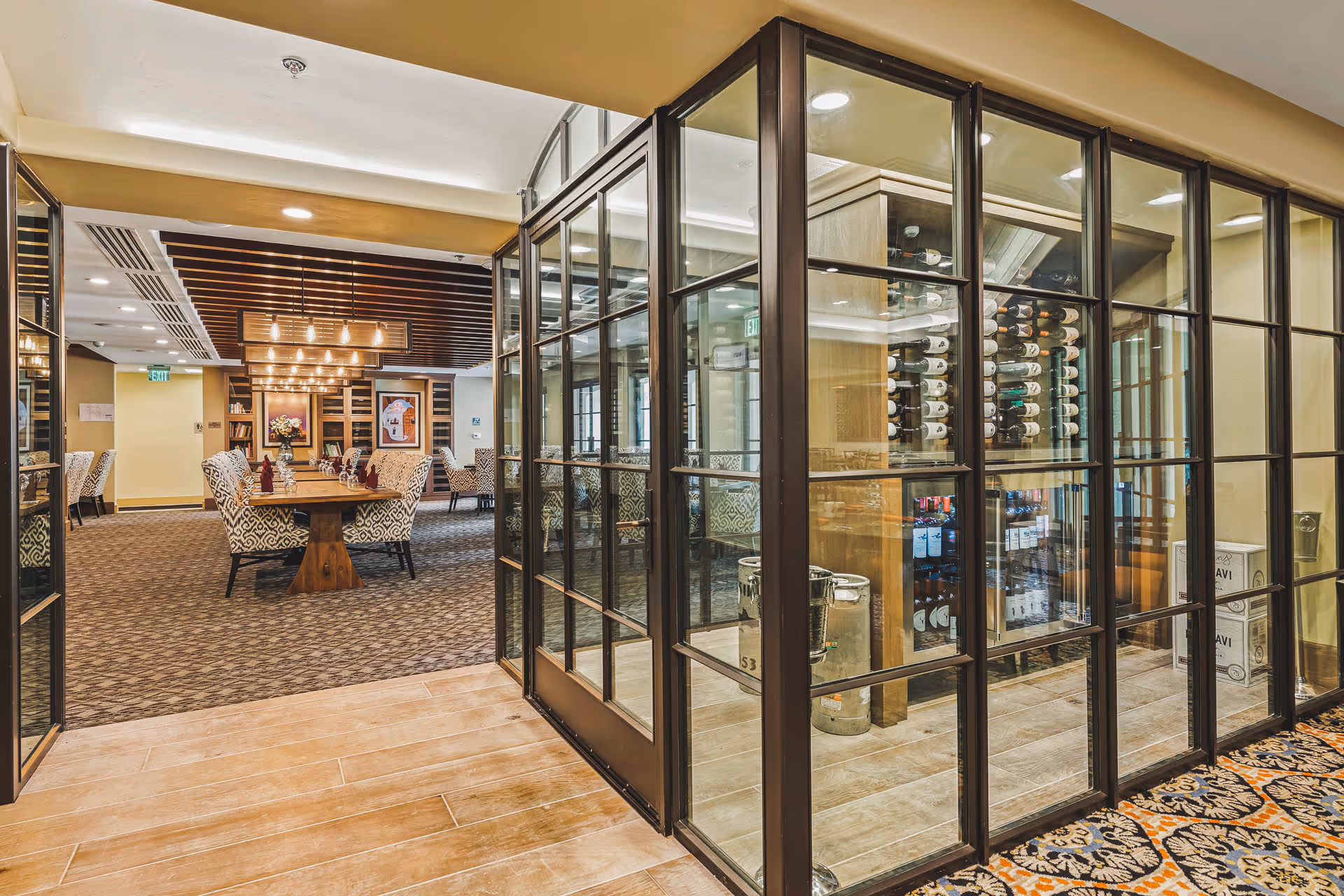Interior view of a dining room in The Hacienda at the Canyon with a wooden table surrounded by patterned upholstered chairs. The room features carpeted flooring, modern pendant lighting, and framed artwork on the walls. Adjacent to the dining area is a glass-enclosed wine storage room with bottles displayed on racks.