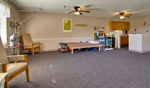 A spacious room with carpeted floor featuring two ceiling fans, several chairs, a treatment table, and medical equipment, with a kitchenette area in the background.