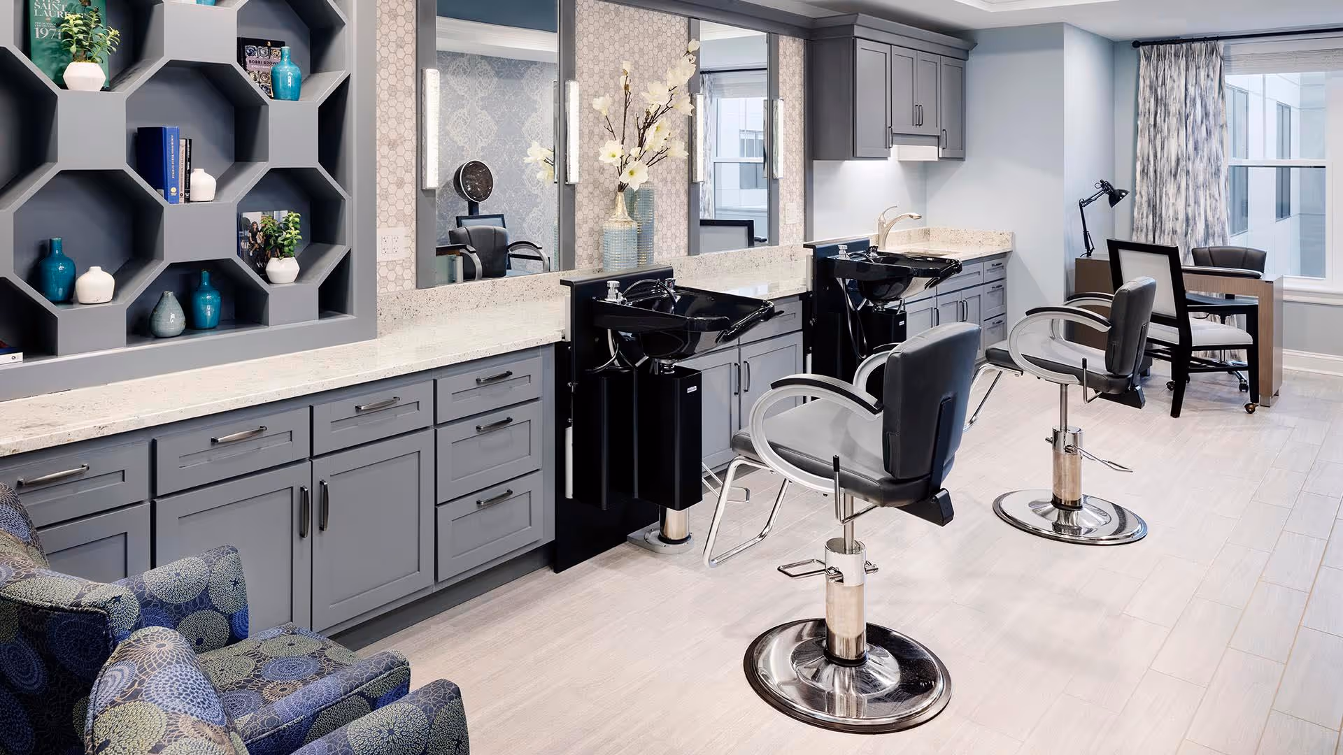 A modern salon area with two black salon chairs in front of black wash basins and grey cabinetry. There is a decorative hexagonal shelf on the left wall with various vases and books. A comfortable patterned armchair is in the foreground, and a desk with a chair and lamp is near a window with curtains in the background.