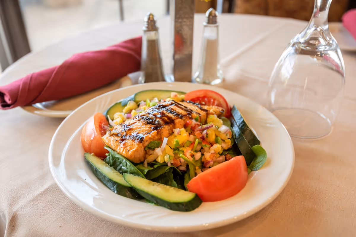 A plate of grilled salmon served on a bed of spinach with a colorful salsa, cucumber slices, and tomato wedges on a white tablecloth. In the background, there is a folded red napkin on a plate, salt and pepper shakers, and an upside-down wine glass.