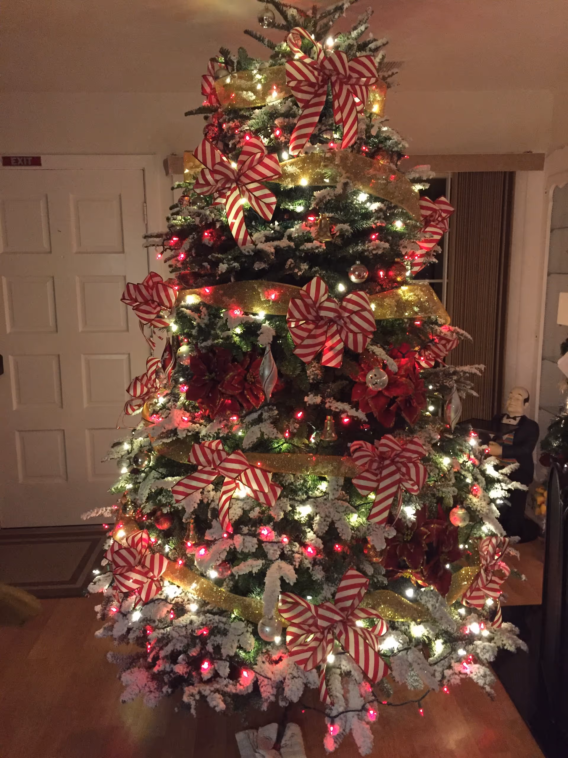 A decorated Christmas tree with red-and-white striped bows, gold ribbon, lights and artificial snow in a living room.