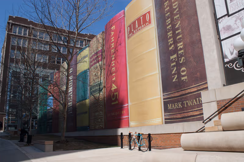 Outdoor view of a building wall decorated with large vertical banners designed to look like the spines of classic books including Fahrenheit 451, Plato The Republic, and Adventures of Huckleberry Finn. There is a bicycle parked near the wall and leafless trees along the sidewalk.