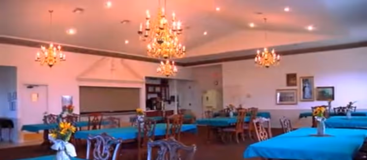 A spacious dining room with multiple tables covered in blue tablecloths, each adorned with a vase of flowers. The room features elegant chandeliers hanging from the ceiling and framed artwork on the walls.