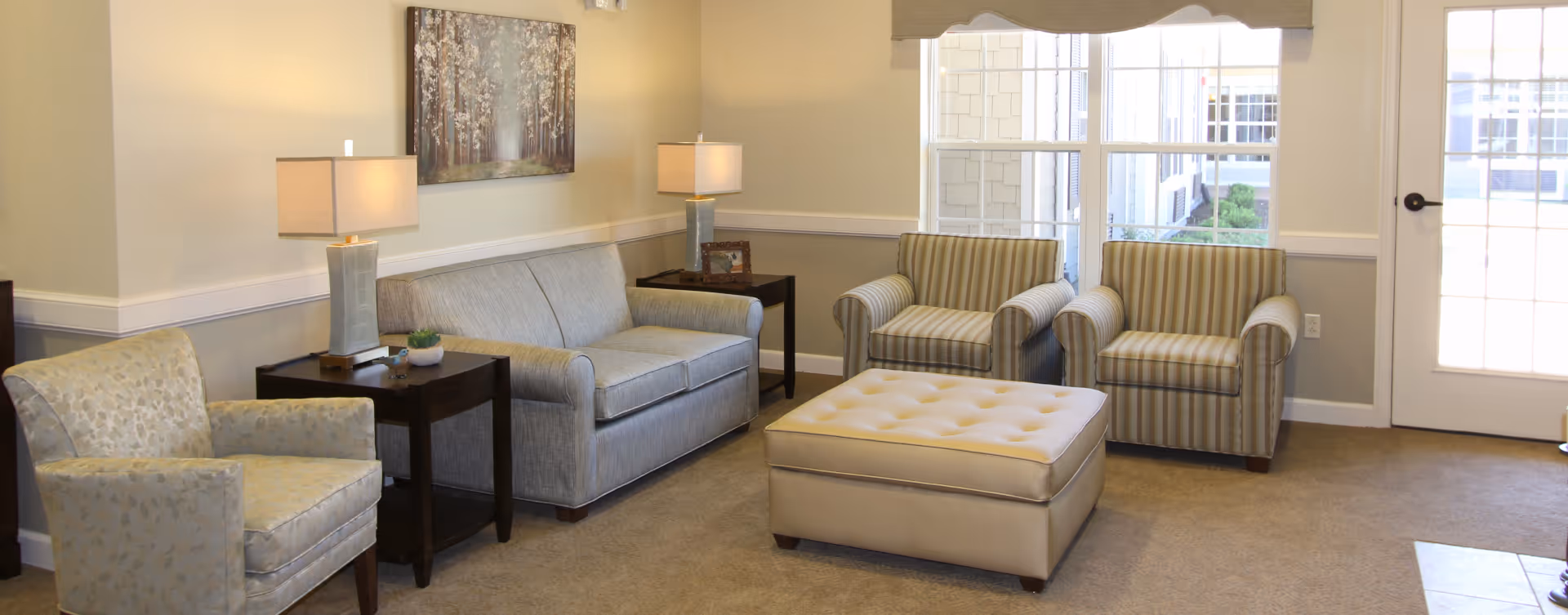 A cozy living room area with a gray sofa, two striped armchairs, a patterned armchair, and a large beige ottoman in the center. There are two side tables with lamps on either side of the sofa, a framed picture on the wall, and a large window and glass door letting in natural light.