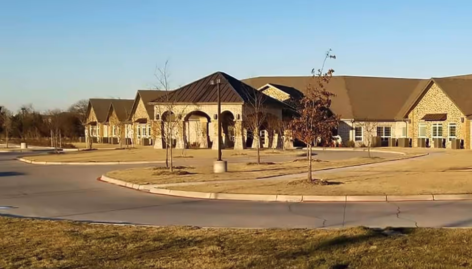 Exterior view of a single-story assisted living facility building with a stone facade, multiple peaked roofs, and a covered entrance. The building is surrounded by a paved driveway, small trees, and a grassy area under a clear sky.