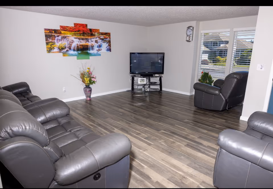 A living room with gray leather sofas and armchairs arranged around a flat-screen TV on a black stand. The room has wood flooring, a wall clock, a large window with white blinds, a colorful waterfall painting on the wall, and a vase with flowers on the floor.