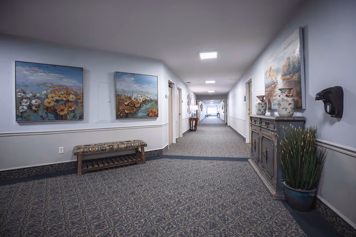 A long, carpeted hallway in a senior living facility with light gray walls and ceiling. The hallway is decorated with framed paintings of flowers and landscapes, a cushioned bench, a wooden cabinet with decorative vases, and a potted plant. Several doors line the hallway, and ceiling lights illuminate the space.
