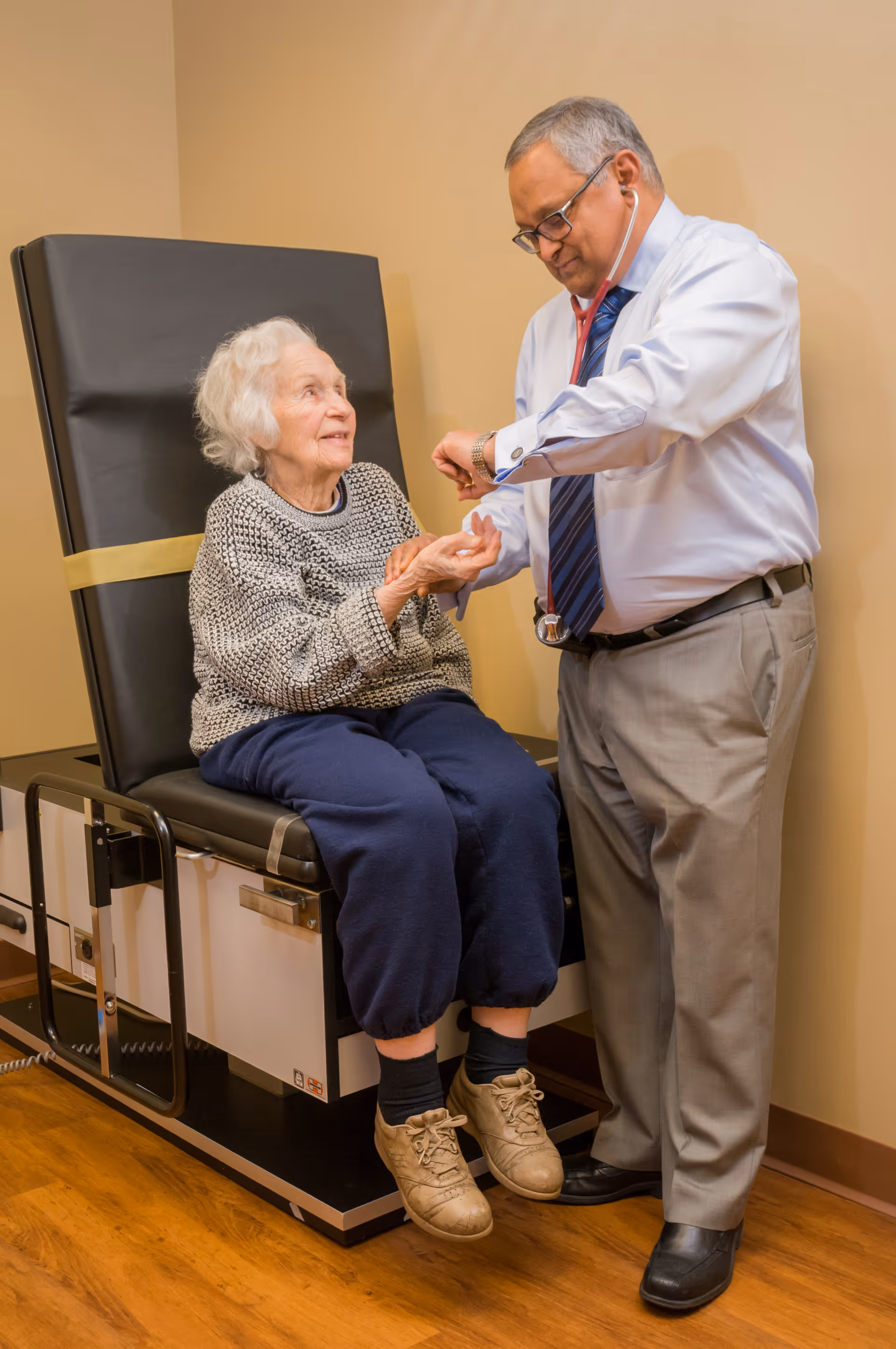 An elderly woman sitting on a medical examination table while a male doctor uses a stethoscope to check her wrist. The woman is smiling and wearing a gray sweater and navy blue pants. The doctor is dressed in a light blue shirt, striped tie, and gray pants.