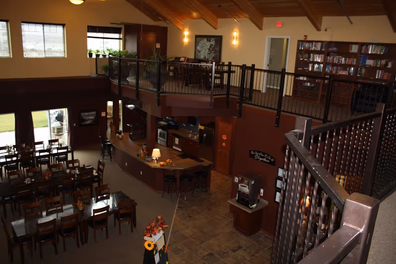 Interior of a communal dining area with multiple tables, a curved service counter/kitchen and an upper mezzanine with bookshelves.