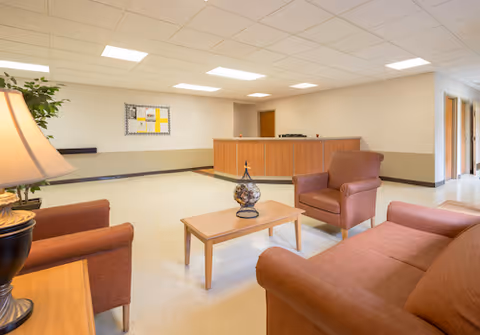 A bright reception lounge with brown upholstered chairs and a sofa around a coffee table facing a wooden reception desk.