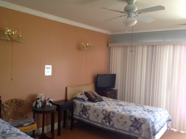 A senior living bedroom with two single beds covered in patterned quilts. There is a small round table with stuffed animals and a chair next to one bed. A small TV sits on a stand in the corner near vertical blinds covering a large window or sliding door. The walls are painted a warm beige color and there are two wall-mounted light fixtures above the beds. A ceiling fan is visible on the white ceiling.