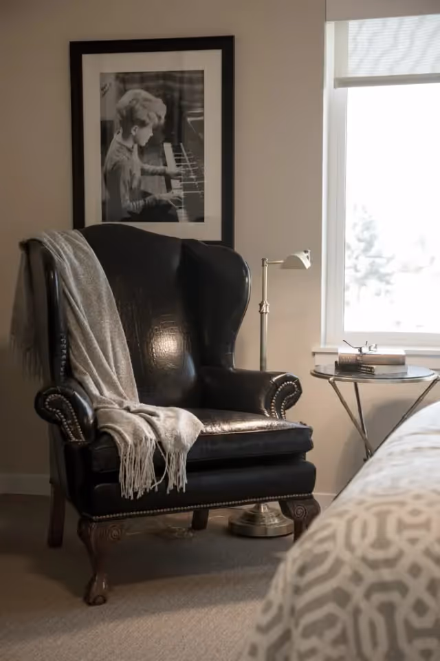 A cozy corner of a bedroom featuring a black leather wingback chair with a gray throw draped over one arm. Next to the chair is a small round side table with books and glasses on it, and a brass floor lamp. A window with a white blind lets in natural light. A framed black and white photograph of a child playing piano hangs on the wall behind the chair.