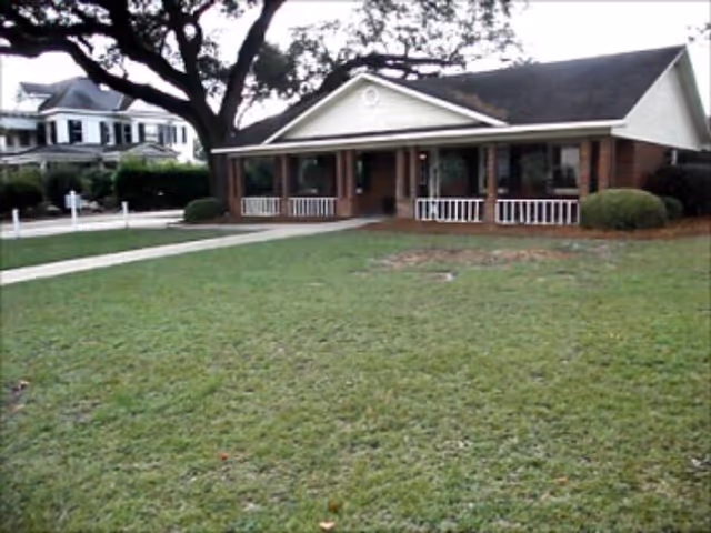 Single-story brick building with a covered porch and white railings, surrounded by a large grassy lawn and a large tree to the left. Another house is visible in the background on the left side.